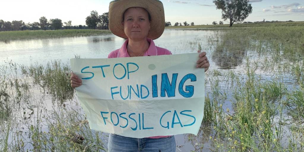Een vrouw staat in een overstroomd weiland. Ze houdt een bord vast met de tekst 'Stop fundING fossil gas'.
