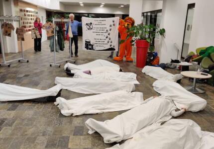 Enkele mensen liggen gewikkeld in witte lakens op de grond alsof ze dood zijn. 